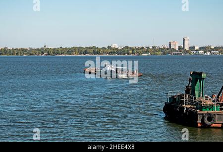 Reinigung der neu entstandenen Insel von Industrieabfällen auf dem Dnjepr mit einem Bagger. Ein Lastkahn mit einem Schlepper nimmt Müll heraus. Ökologisches Konzept. Stockfoto