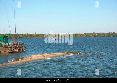 Reinigung der neu entstandenen Insel von Industrieabfällen auf dem Dnjepr mit einem Bagger. Ein Lastkahn mit einem Schlepper nimmt Müll heraus. Ökologisches Konzept. Stockfoto