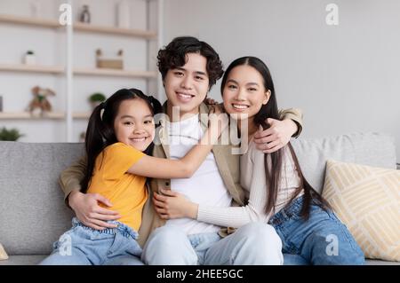 Fröhliche asiatische Paar und Tochter Umarmungen sitzt auf dem Sofa im Wohnzimmer Interieur Stockfoto