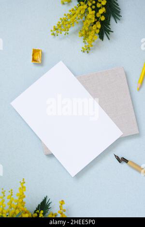 Weißes Blatt Papier für Textkopien, umgeben von Frühlingsmimosen, Notizblock, gelbem Bleistift, Wasserfarben in Küvetten auf blauem Hintergrund. Arbeitsplatz Stockfoto