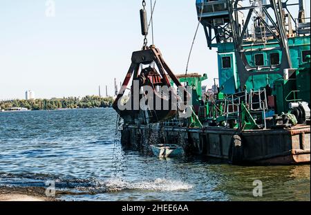 Reinigung der neu entstandenen Insel von Industrieabfällen auf dem Dnjepr mit einem Bagger. Der Eimer lädt den Schlamm auf den Lastkahn. Ökologisches Konzept. Stockfoto