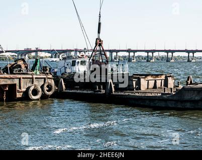 Reinigung der neu entstandenen Insel von Industrieabfällen auf dem Dnjepr mit einem Bagger. Der Eimer lädt den Schlamm auf den Lastkahn. Ökologisches Konzept. Stockfoto