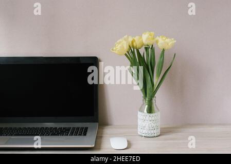 Frühling Home Office Stillleben Komposition. Leerer Laptop-Bildschirm. Gelbe Tulpen in einer Vase auf dem Tisch. Büroarbeitsplatz. Skandinavische Innenausstattung. Stockfoto