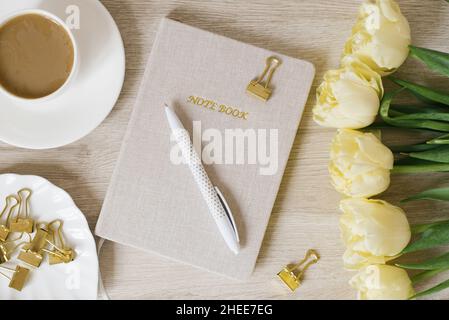 Frühstückskaffeebecher, Notizblock, Stift, Büroklammern auf einem Teller und gelbe Tulpen-Blumen auf einem Holztisch im flachen Lay-Stil. Schreibtisch für Frauen. Stockfoto