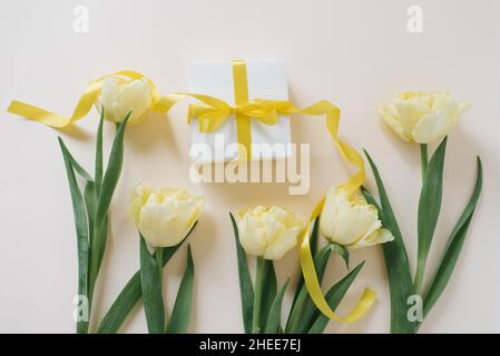 Geschenkschachtel in weißem Papier mit einem gelben Band und Blumen aus gelben Tulpen auf einem hellen Tisch verpackt. Flach liegend. Stockfoto