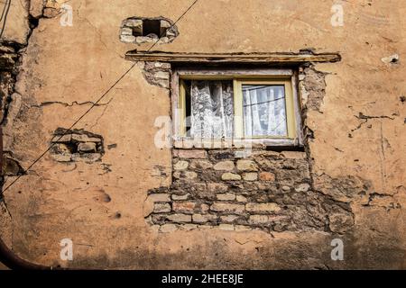 Fenster im oberen Stockwerk mit hübschen Spitzvorhängen und eine Seite offen in sehr grungiger und bröckelnder Wand mit einer diagonalen Versorgungslinie - die Wand ist rau und texturiert Stockfoto