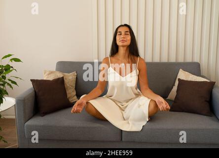 Junge friedvolle Frau, die mit geschlossenen Augen in Lotusposition auf dem Sofa zu Hause sitzt und am Morgen meditiert Stockfoto