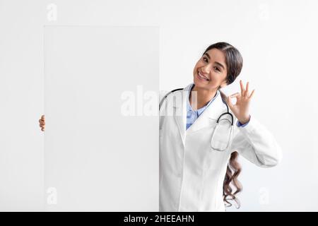 Froh junge nette indische Dame Arzt in Uniform zeigt ok Zeichen mit Hand hält große Banner mit Leerzeichen Stockfoto