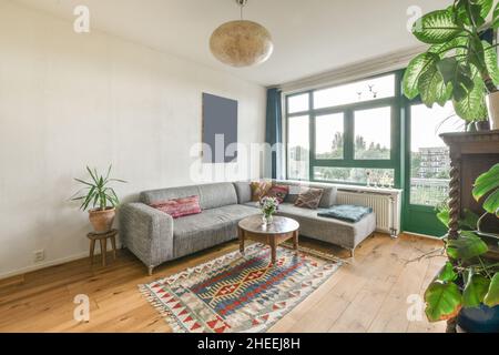 Komfortables Ecksofa mit dekorativen Kissen und kleinem runden Couchtisch im eklektischen Wohnzimmer mit Holzmöbeln und großem Fenster Stockfoto