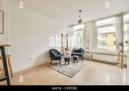 Die Einrichtung des geräumigen, hellen Wohnzimmers mit kleinen Stühlen und bequemen Sesseln neben dem Tisch, die auf einem Teppich in der Nähe der Fenster platziert sind Stockfoto