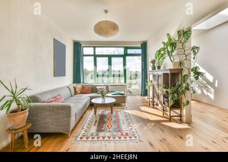 Komfortables Ecksofa mit dekorativen Kissen und kleinem runden Couchtisch im eklektischen Wohnzimmer mit Holzmöbeln und großem Fenster Stockfoto