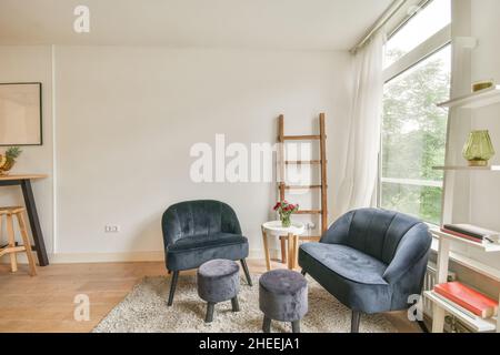 Die Einrichtung des geräumigen, hellen Wohnzimmers mit kleinen Stühlen und bequemen Sesseln neben dem Tisch, die auf einem Teppich in der Nähe der Fenster platziert sind Stockfoto