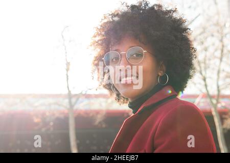 Positive schwarze Hündin mit Afro-Frisur und Brillen in purpurrotem Mantel und Blick auf die Kamera im Sonnenlicht Stockfoto