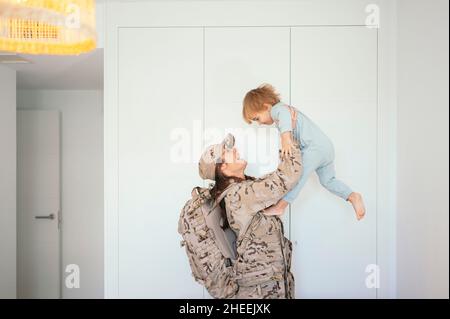 Seitenansicht einer glücklichen Soldatin in Uniform mit Rucksack, der einen niedlichen Sohn in ausgestreckten Armen in der Nähe einer Holzkrippe im Schlafzimmer hält Stockfoto