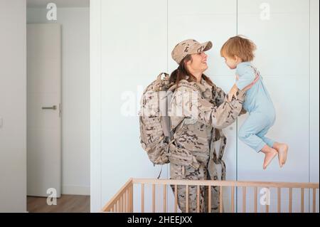 Seitenansicht einer glücklichen Soldatin in Uniform mit Rucksack, der einen niedlichen Sohn in ausgestreckten Armen in der Nähe einer Holzkrippe im Schlafzimmer hält Stockfoto