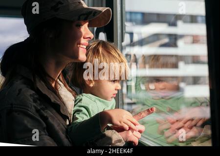 Seitenansicht einer lächelnden Soldatin in Uniform, die aus dem Fenster mit einem niedlichen Sohn blickt, während sie im hellen Raum mit Sonnenlicht steht Stockfoto