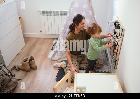 Seitenansicht einer positiven Militärmutter in Uniform, die neben dem Sohn steht und mit einem vielbeschäftigten Brett im Spielzimmer mit einer Decke spielt Stockfoto