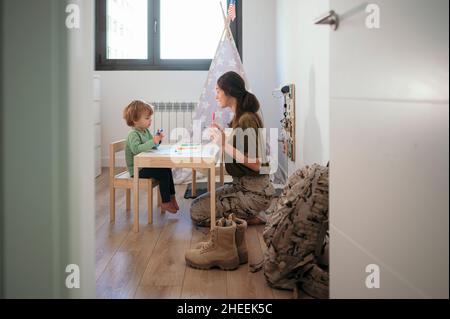 Seitenansicht der fürsorglichen Militärmutter in Uniform und der Zeichnung des Sohnes mit bunten Filzstiften auf Papier im hellen Raum mit Camouflage-Rucksack Stockfoto