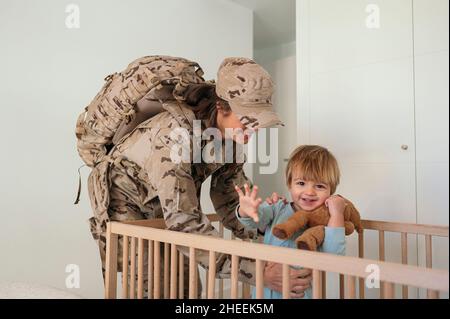 Seitenansicht der glücklichen weiblichen Soldaten in Uniform mit Rucksack hält niedlichen Sohn in Holzkrippe im Schlafzimmer Stockfoto
