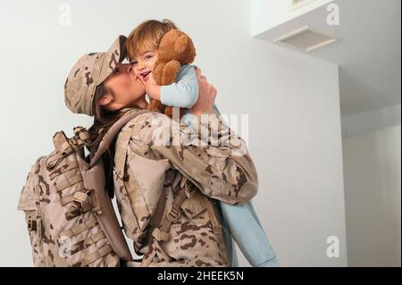 Seitenansicht der glücklichen weiblichen Soldaten in Uniform mit Rucksack, der küssenden niedlichen Sohn im Schlafzimmer hält Stockfoto