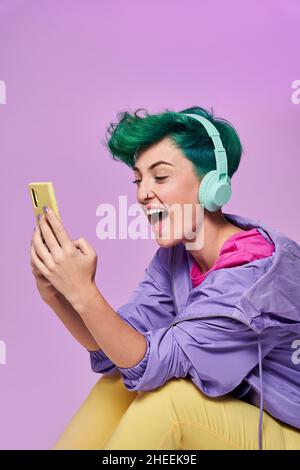 Fröhliche Hündin mit Kopfhörern im Retro 80s Tragen Sie im Studio beim Surfen in sozialen Medien auf dem Handy auf lila Stockfoto