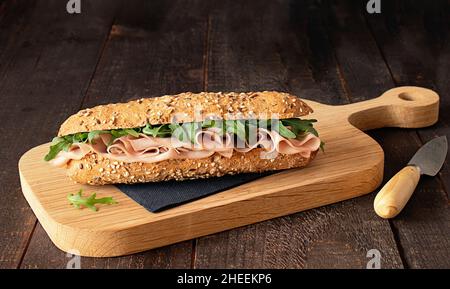 Mortadella-Schinken-Sandwich mit Rucola-Blättern auf Holzschneidebrett auf rustikalem Küchentisch Stockfoto