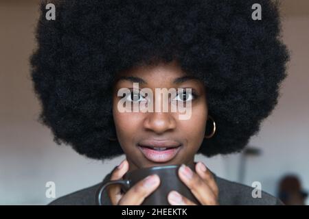 Lächelnde afroamerikanische Frau mit Afro-Frisur und einer Tasse Kaffee in den Händen, die im hellen Raum stehen und die Kamera betrachten Stockfoto