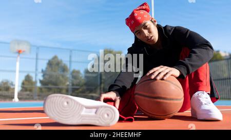 Seriöser asiatischer Mann in Sportkleidung mit Basketball in der Hand, der während des Trainings auf dem Sportplatz sitzt und die Kamera anschaut Stockfoto