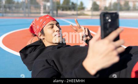 Inhalt sportlicher asiatischer Mann zeigt V-Zeichen und nimmt Selbstporträt auf dem Smartphone auf, während er während des Trainings auf dem Sportplatz liegt Stockfoto