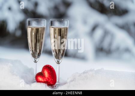 Nahaufnahme von zwei Sektflöten, die im Schnee kühlen und im roten Herzen auf dem Hintergrund der Winterlandschaft. Konzept für die Valentinstag-Feier. Speicherplatz kopieren. Stockfoto
