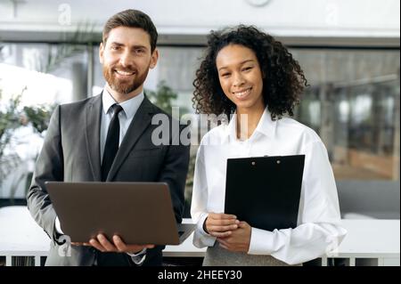 Zwei erfolgreiche Geschäftsleute, kaukasischer Mann ceo des Unternehmens, afroamerikanische weibliche Top-Managerin, in formeller Kleidung, stehen im modernen Büro, schauen auf die Kamera, lächelnd, Partnerschaftskonzept Stockfoto