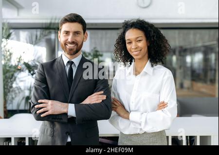 Einflussreiche, erfolgreiche Mitarbeiter verschiedener Nationalitäten, kaukasischer Mann und afroamerikanische Frau, in formeller Kleidung, stehen mit gekreuzten Armen im modernen Büro, blicken auf die Kamera und lächeln Stockfoto