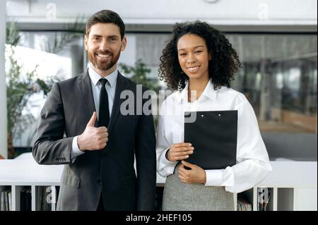 Positive erfolgreiche multirassische Geschäftspartner, afroamerikanische Geschäftsfrau, kaukasischer Geschäftsmann, Firmenangestellte, die in einem modernen Büro stehen, auf die Kamera schauen und freundlich lächeln Stockfoto