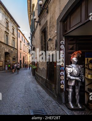 Orvieto, Umbrien, 2021. August – Fußgängerzone im historischen Zentrum von Orvieto mit einer mittelalterlichen Rüstung, die in einem Souvenirladen ausgestellt ist. Stockfoto