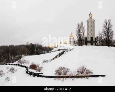 Blick auf das National Museum of Holodomor Denkmal für Völkermord oder Hungeropfer und die goldenen Kuppeln von Kiev Pechersk Lavra oder das Kiewer Höhlenkloster auf schneebedecktem Schnee Stockfoto