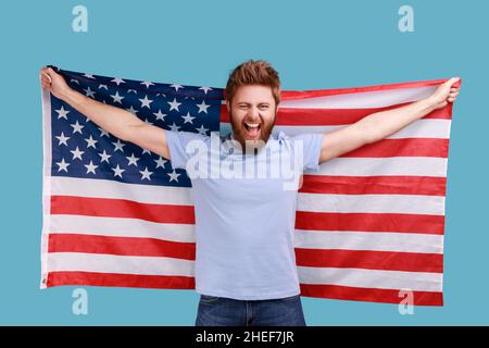 Porträt eines hübschen, überfröhlichen bärtigen Mannes, der die US-Flagge hält und mit frohlockender Anschauung auf die Kamera blickt und Nationalfeiertage feiert. Innenaufnahme des Studios isoliert auf blauem Hintergrund. Stockfoto