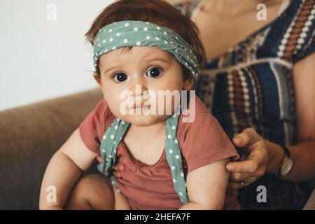 Porträt eines entzückenden Babys, das an den Händen der Großmutter hält und ein Stirnband trägt und direkt auf die Kamera blickt. Schönes junges Mädchen. Stockfoto