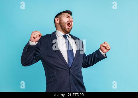 Junger attraktiver Geschäftsmann mit Bart in einem dunklen Anzug im offiziellen Stil, der gähnt und die Hände hochhebt und sich von Überstunden ermüdet fühlt Innenaufnahme des Studios auf blauem Hintergrund. Stockfoto