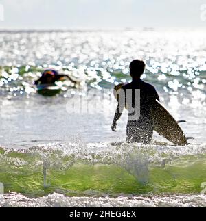 Ein silhouettierter Surfer, der am Kuta Beach auf der Insel Bali in Indonesien in Richtung Brandung geht. Stockfoto