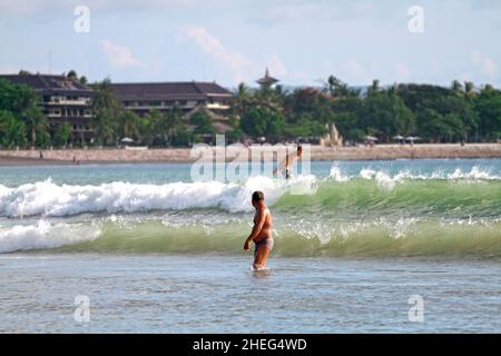 Ein kaukasischer Surfer reitet auf einer Welle mit einem einheimischen Balinesen im Vordergrund am Kuta Beach auf der Insel Bali in Indonesien. Stockfoto