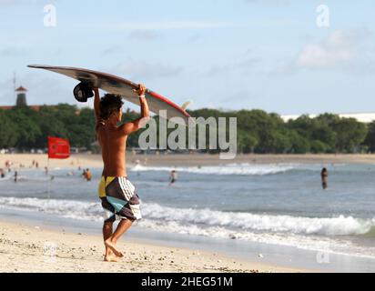 Ein junger balinesischer Surfer, der sein Surfbrett auf dem Kopf am Kuta Beach auf der Insel Bali in Indonesien trägt. Stockfoto