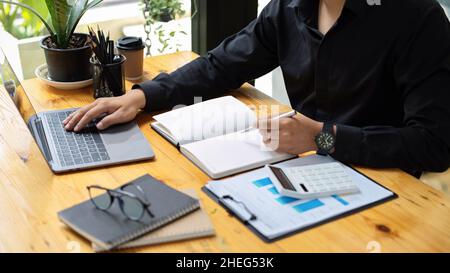 Männlicher Buchhalter, der seine Geschäftsaufgabe im Büro macht. Geschäftsmann, der am Laptop arbeitet. Bericht und Rechner auf dem Tisch. Stockfoto