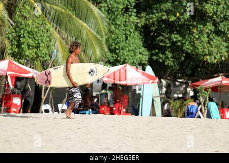 Ein junger Indonesier, der am Kuta Beach auf der Insel Bali in Indonesien den Strand entlang läuft und ein Surboard trägt. Stockfoto