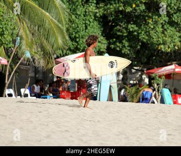 Ein junger Indonesier, der am Kuta Beach auf der Insel Bali in Indonesien den Strand entlang läuft und ein Surboard trägt. Stockfoto