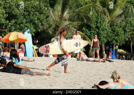 Ein junger Indonesier, der am Kuta Beach auf der Insel Bali in Indonesien den Strand entlang läuft und ein Surboard trägt. Stockfoto