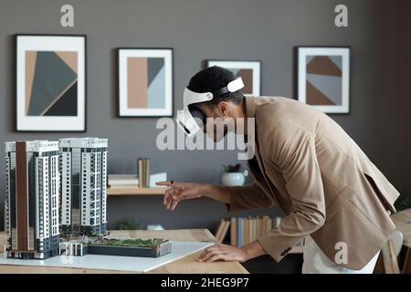 Junger biracial Geschäftsmann in vr Headset Biegen über Tisch und zeigt auf Haus-Layout, während die virtuelle Präsentation Stockfoto