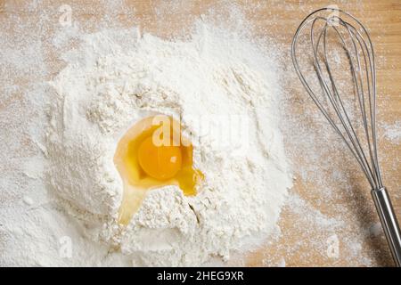 Flatlay von gebrochenem rohem Ei auf den Haufen Mehl für die Herstellung von hausgemachtem Teig vorbereitet und Schneebesen auf Holztisch Stockfoto
