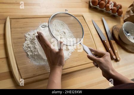 Die Hände der Küchenchefin sieben Mehl über dem Schneidebrett am hölzernen Küchentisch mit Eiern, Messern und einer Schüssel Stockfoto