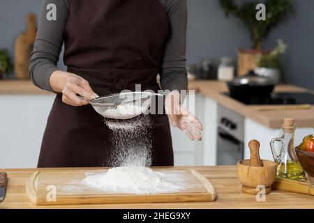 Zeitgenössische Frau in der Schürze sieben Mehl am hölzernen Küchentisch, während sie Zutaten für hausgemachten Teig und Gebäck zubereitet Stockfoto