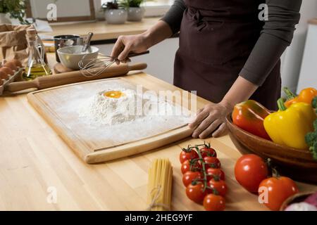 Junge Frau in der Schürze hält Schneebesen über den Haufen Mehl mit rohem Ei auf der Oberseite, während Sie hausgemachten Teig für Gebäck zubereiten Stockfoto